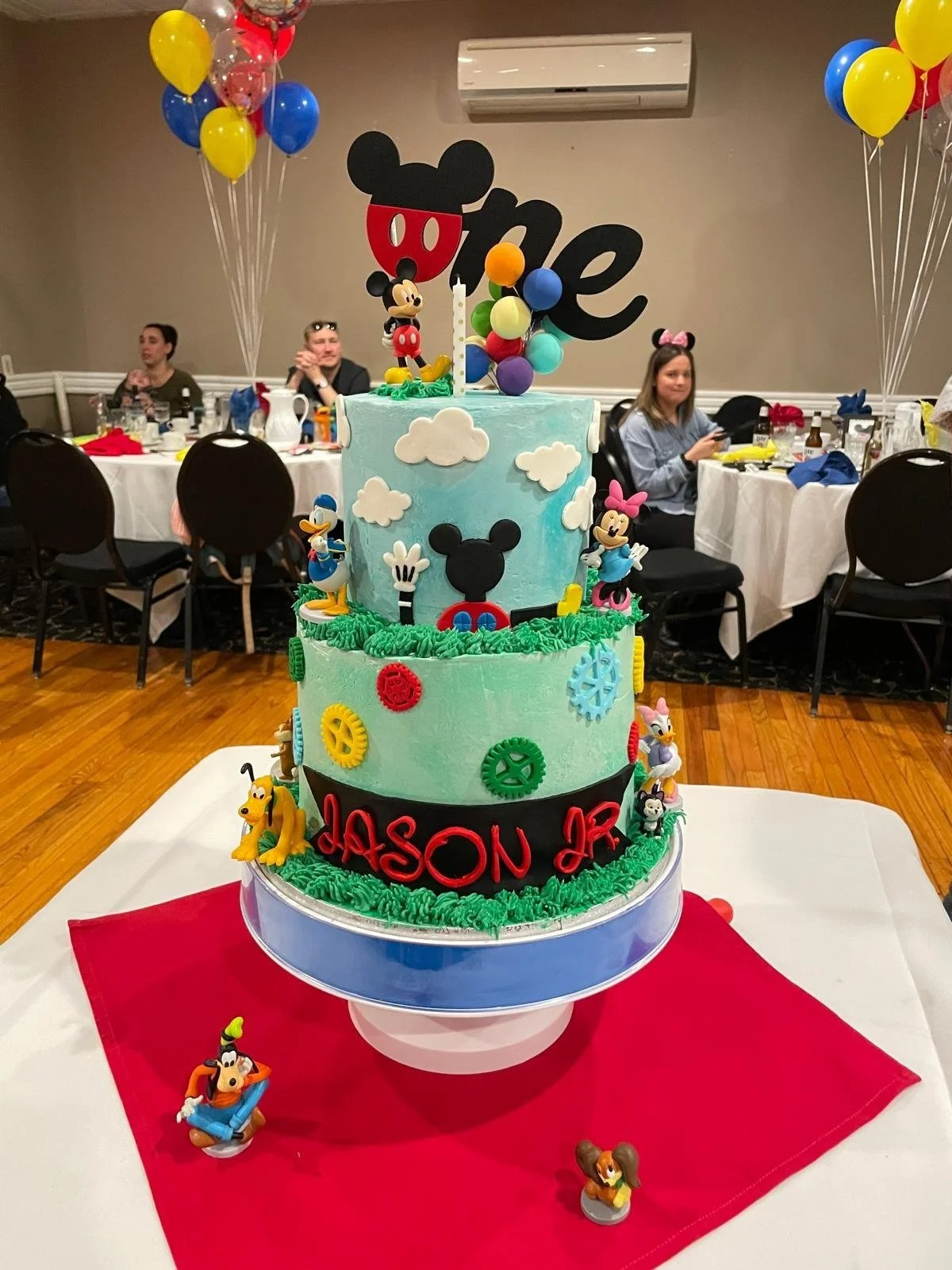 A colorful three-tiered birthday cake decorated with Disney character toppers, including Mickey Mouse, Minnie Mouse, Donald Duck, Goofy, and Daisy Duck. The cake features clouds and gears, with a birthday message in red lettering that says 'JASON JR'. Balloons in yellow, blue, and red are attached to the cake. The cake is on a white table with a red tablecloth, and a party room with guests, tables, and chairs in the background.