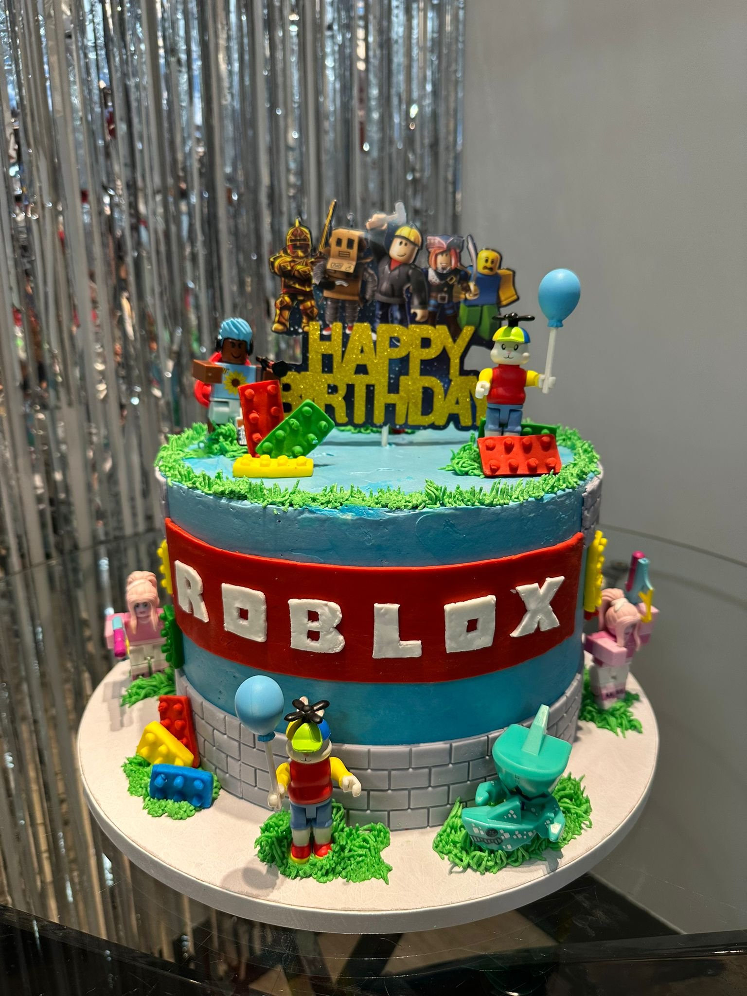 A two-tiered birthday cake decorated with Roblox and Minecraft themes, featuring figurines, a "Happy Birthday" sign, and colorful decorations, set against a background of silver streamers.