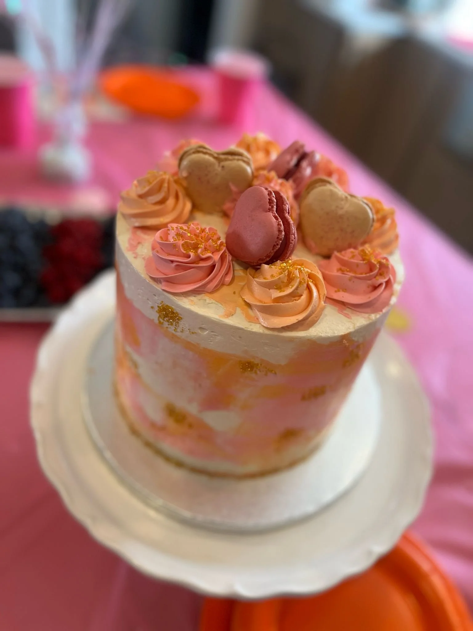 A cake decorated with pink and peach-colored frosting swirls, macarons, and heart-shaped cookies on top.