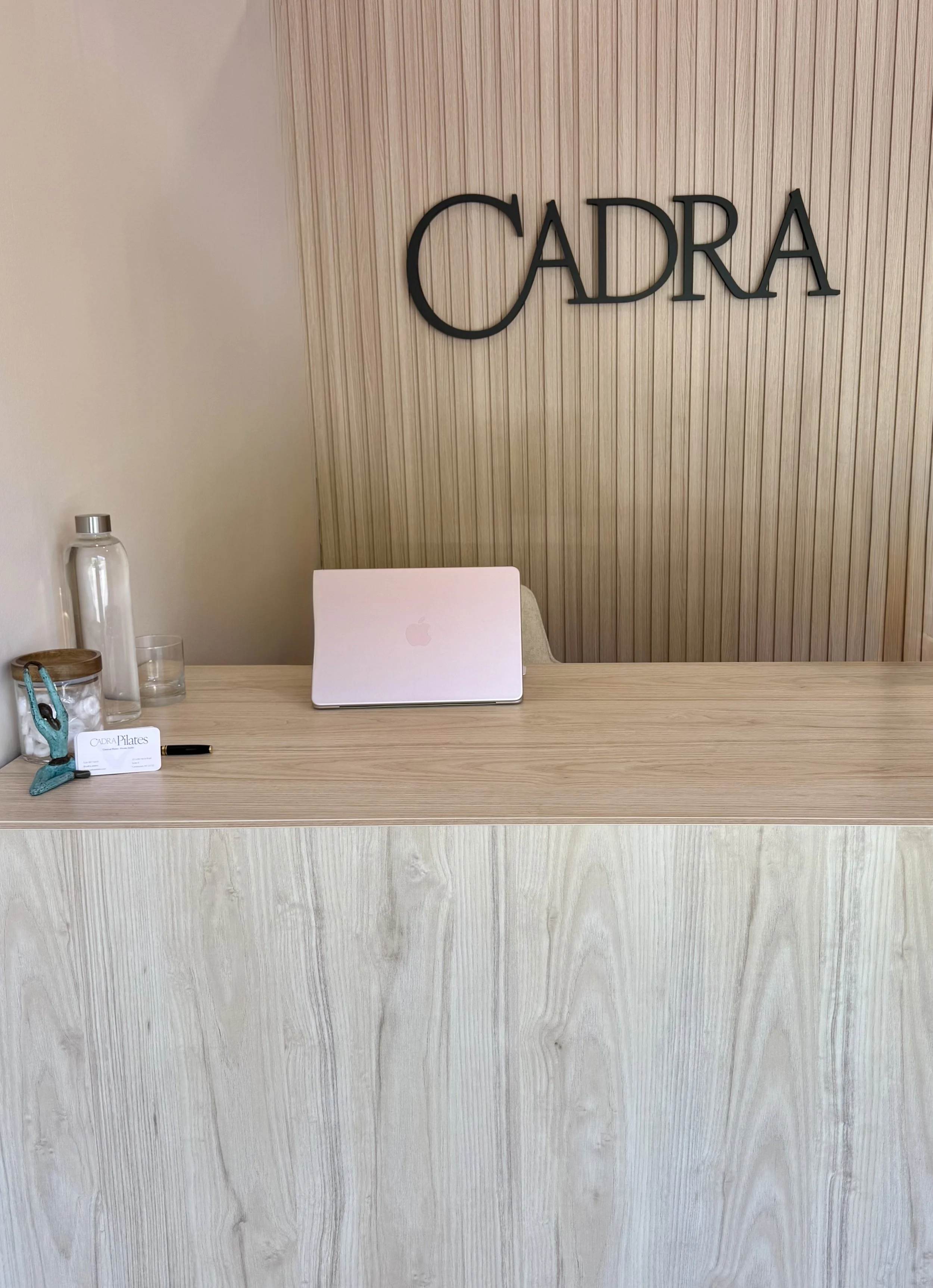 Reception desk with a closed silver laptop, a business card with 'Cadra Pilates' logo, a model yoga pose sculpture, a water bottle, and a drinking glass. The background has a wooden textured wall with 'Cadra' written in large black letters.