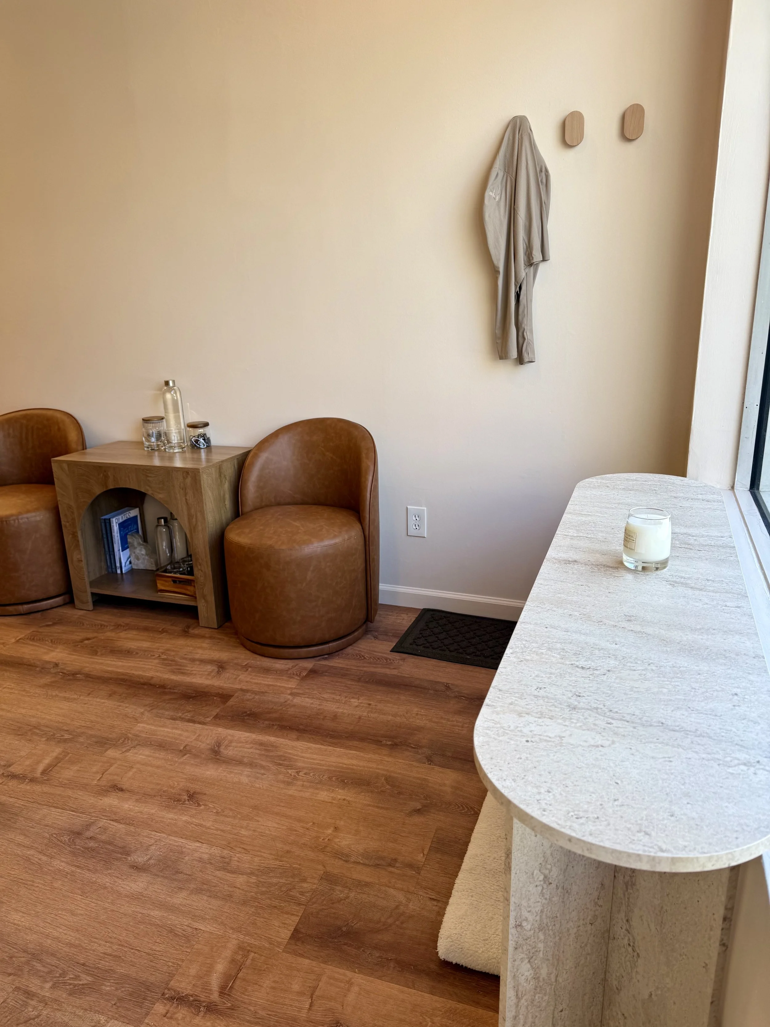 A corner of a cozy room with wooden flooring, a small wooden table with glass bottles and books, two brown leather chairs, a beige wall with two decorative wooden pieces, a shirt hanging on the wall, and a table with a candle near a window.