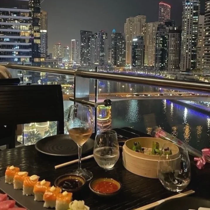 A nighttime cityscape view from a balcony restaurant, with a table set for dining that includes sushi, wine, water, and dipping sauces, overlooking a river and illuminated skyscrapers in the background.