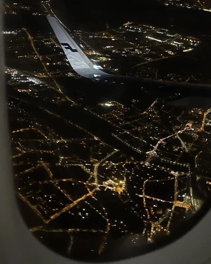 Nighttime aerial view of a city from an airplane window showing illuminated streets and buildings.