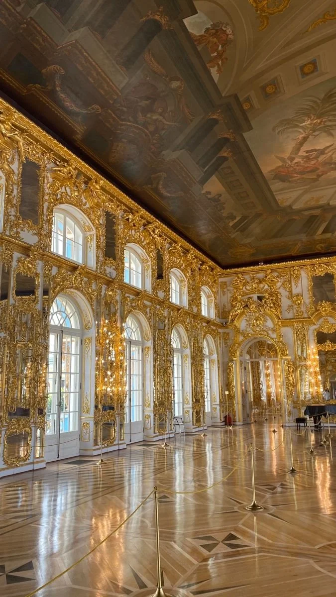 Luxurious gold-embellished room with tall arched windows, an ornate ceiling, and a polished wooden floor, likely part of a palace or historic building.