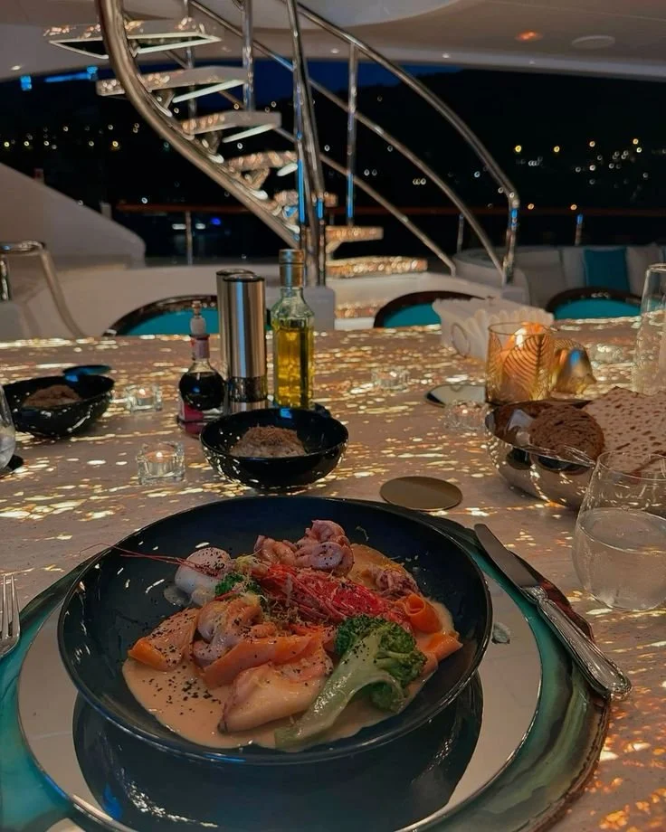 A restaurant table with a seafood dish featuring shrimp, vegetables, and sauce in a black bowl, set with utensils, glassware, condiments, and decorative lighting in the background.