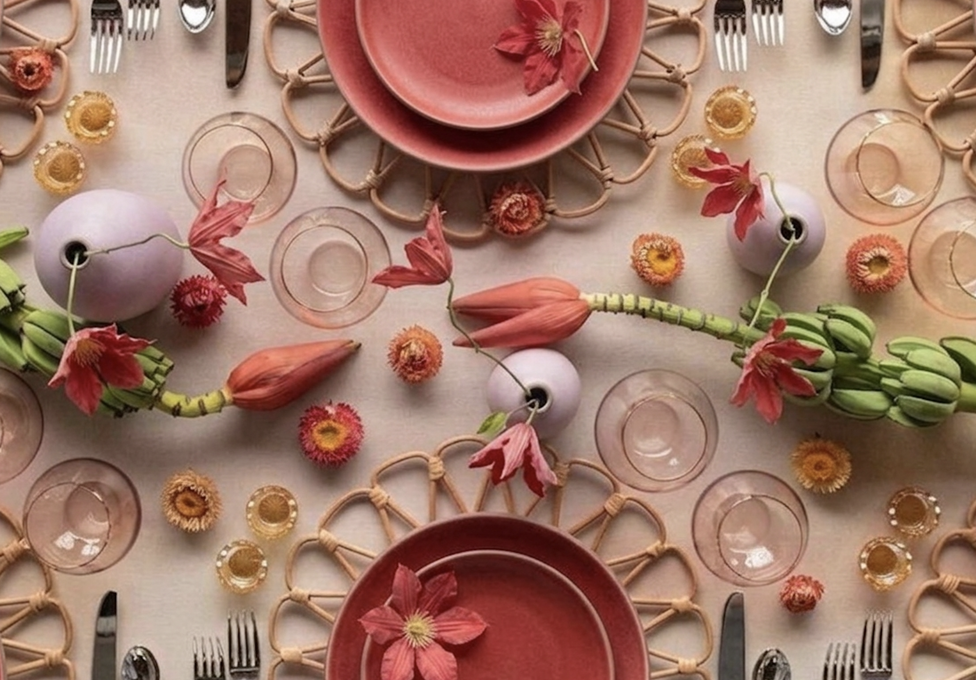 Elegant table setting with pink plates, glassware, cutlery, and floral decorations including large pink and orange flower buds and smaller colorful flowers, arranged on a light-colored tablecloth.
