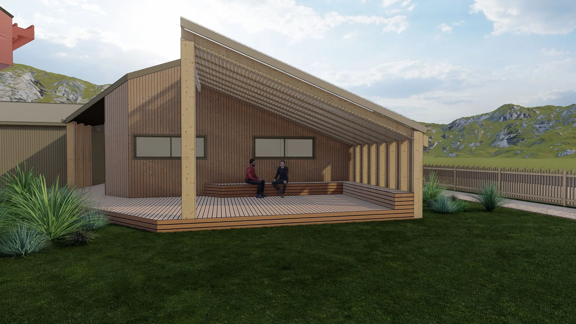 A wooden outdoor pavilion with a sloped roof and seating area, with two people sitting and talking, surrounded by grass, plants, and a wooden fence, with hills in the background.
