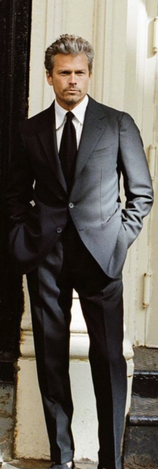 Man with gray hair dressed in a black suit, white shirt, and black tie, standing outside against a doorframe.