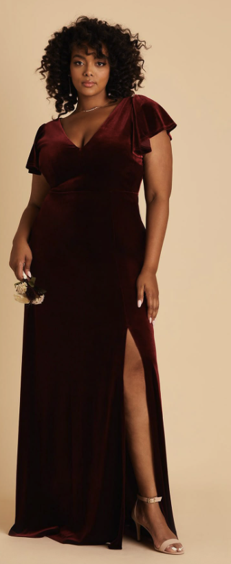 Woman wearing a long, burgundy velvet gown with a thigh-high slit and ruffled sleeves, standing against a neutral background.