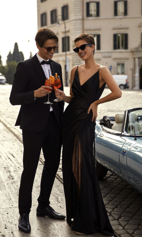 A man in a tuxedo and a woman in a black evening gown, both wearing sunglasses, stand outside on a cobblestone street near a vintage convertible car. They are smiling, holding glasses of red cocktails, and appear to be enjoying a festive night out.