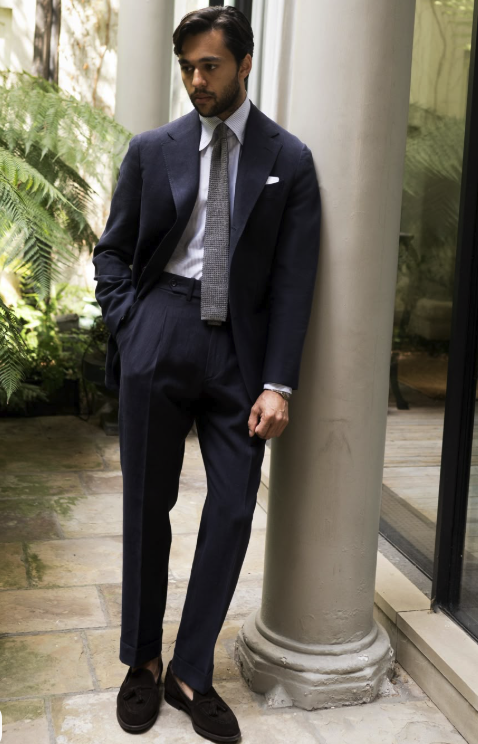 Man in a dark blue suit, white shirt, and gray tie leaning against a column in a glass-enclosed area with greenery outside.