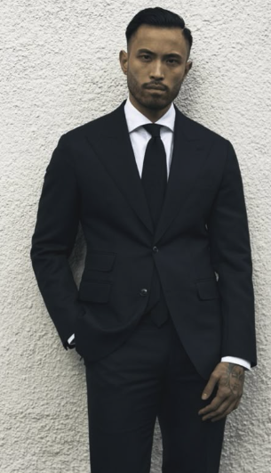 A man wearing a black suit and tie, standing against a textured white wall, with a serious expression.