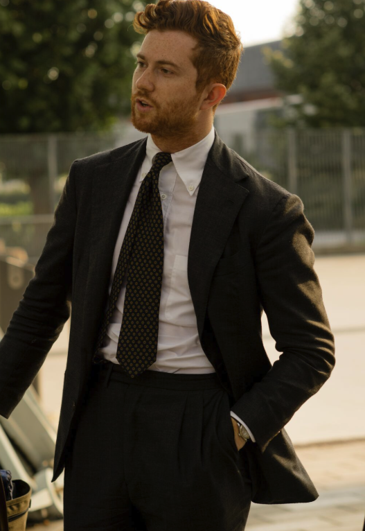 A man with red hair and a beard wearing a dark suit, white shirt, and patterned tie, standing outdoors with a fence and trees in the background.