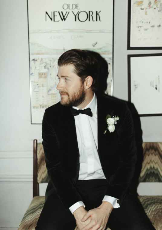 A man with a beard and hairstyle in a tuxedo with a bow tie and boutonniere, sitting on a vintage-style chair against a backdrop of framed maps and artwork.