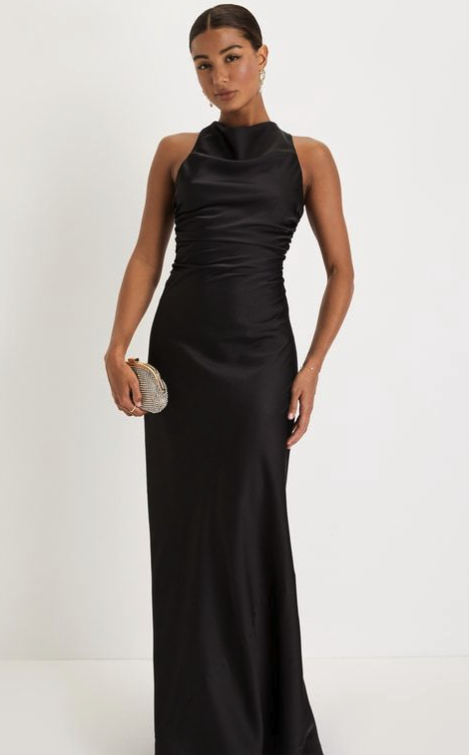 A woman in a sleeveless, high-neck black satin gown, holding a small silver clutch, standing against a plain white background.