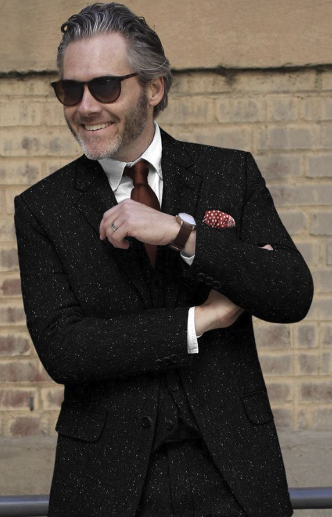 A man with gray hair and beard, wearing sunglasses, a black glittery suit, white shirt, maroon tie, and a red polka dot pocket square, standing in front of a brick wall.