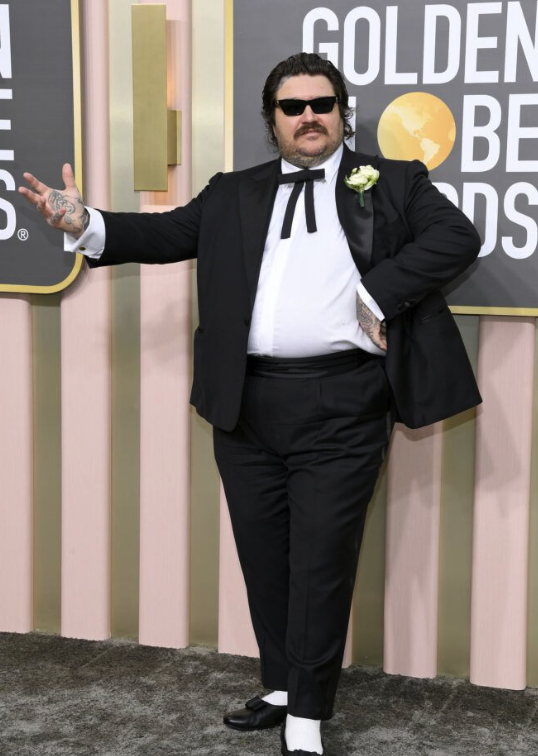 A man with dark hair, mustache, and sunglasses wearing a black tuxedo with a white shirt and bow tie, standing at the Golden Globe Awards, posing with one hand extended and the other on his waist.