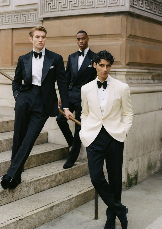 Three men dressed in formal tuxedos posing on steps outside a building with ornate architectural details.