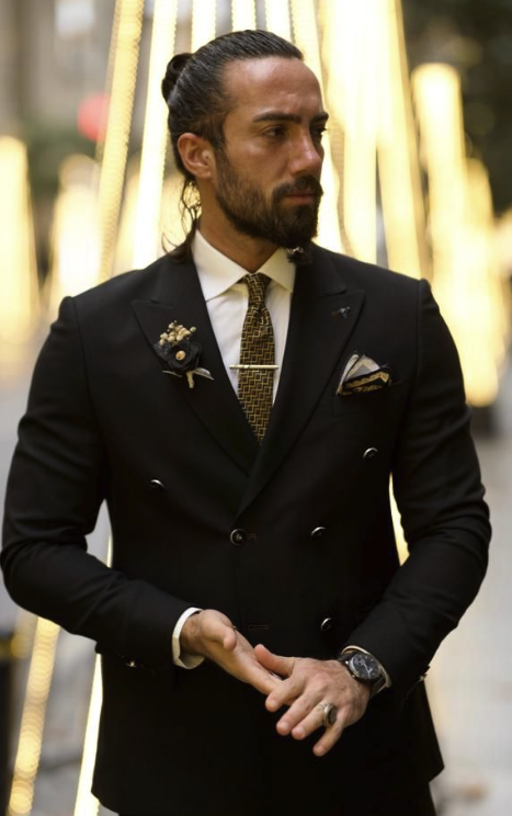 A man with long dark hair and a beard, wearing a black double-breasted suit with a white shirt, patterned tie, and pocket square. He is standing in front of a backdrop of vertical illuminated lights, with his hands clasped in front of him, wearing a