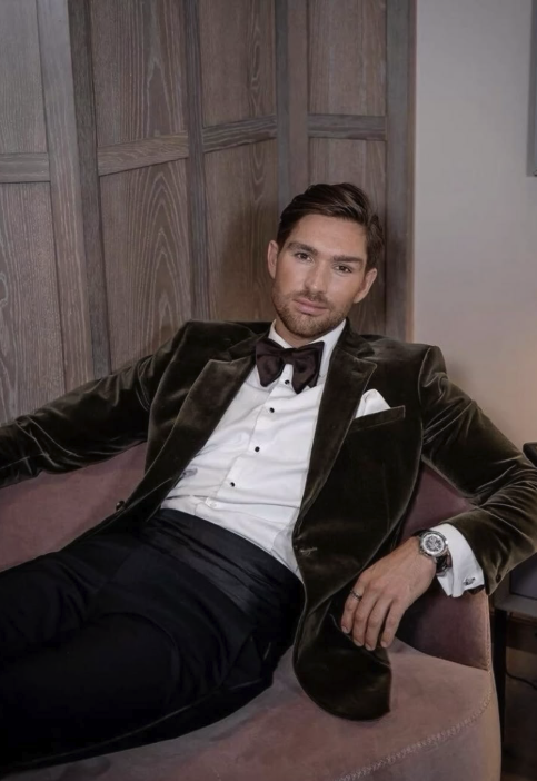 A man sitting on a sofa, wearing a tuxedo with a velvet blazer, white dress shirt, black bow tie, and a watch, with wooden paneling behind him.