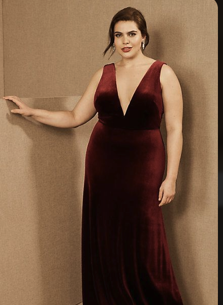 A woman in a long, deep red velvet evening gown with a plunging neckline, standing against a beige background.