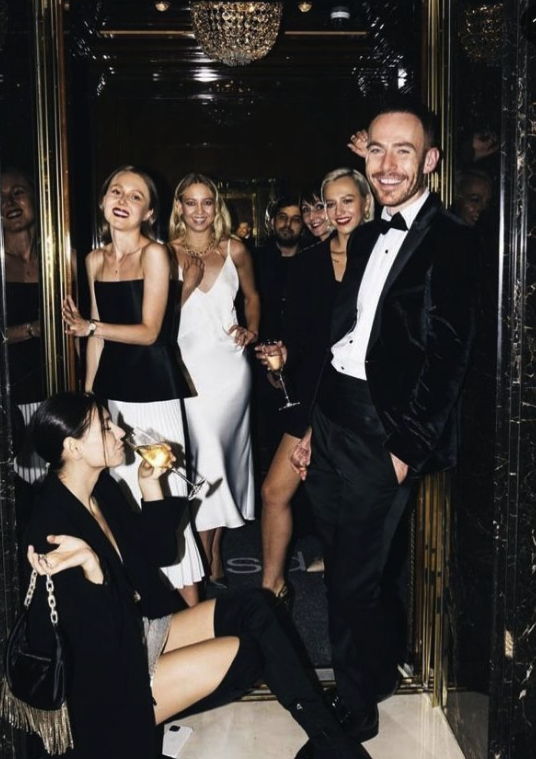 Group of elegantly dressed people at a party with a woman sitting on the floor drinking from a glass, a man in a tuxedo smiling at the camera, and other women in stylish dresses inside a luxurious venue.