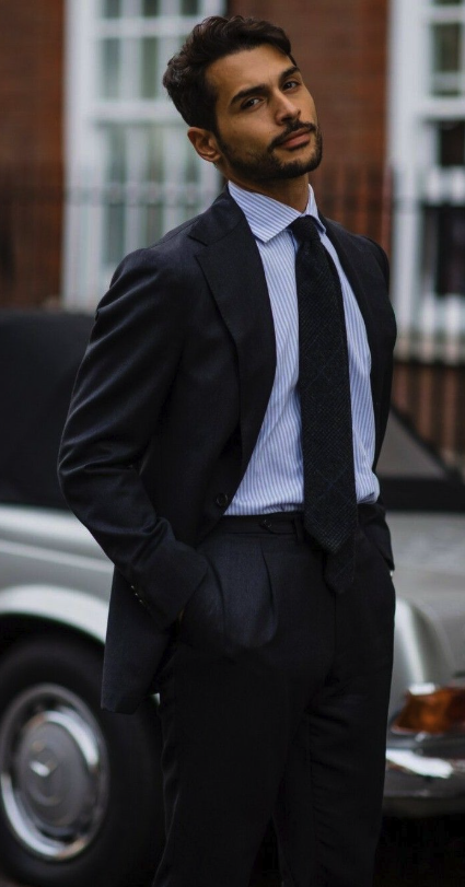 A young man with dark hair and a beard in a dark suit and tie standing outdoors in front of a black car and brick building.