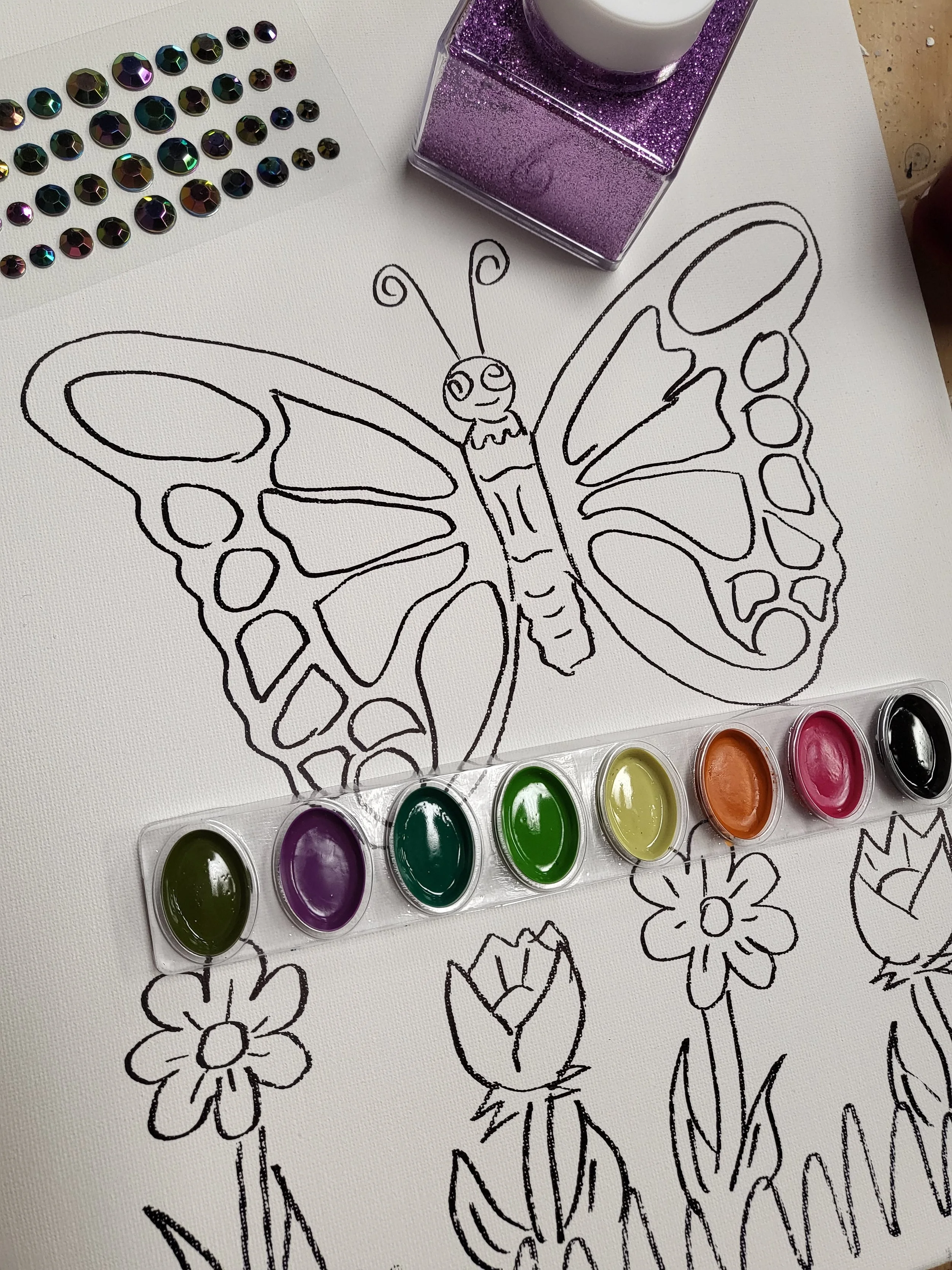 A line drawing of a butterfly on white paper with flowers, surrounded by watercolor paint pots, rhinestones, and a purple glitter candle holder.