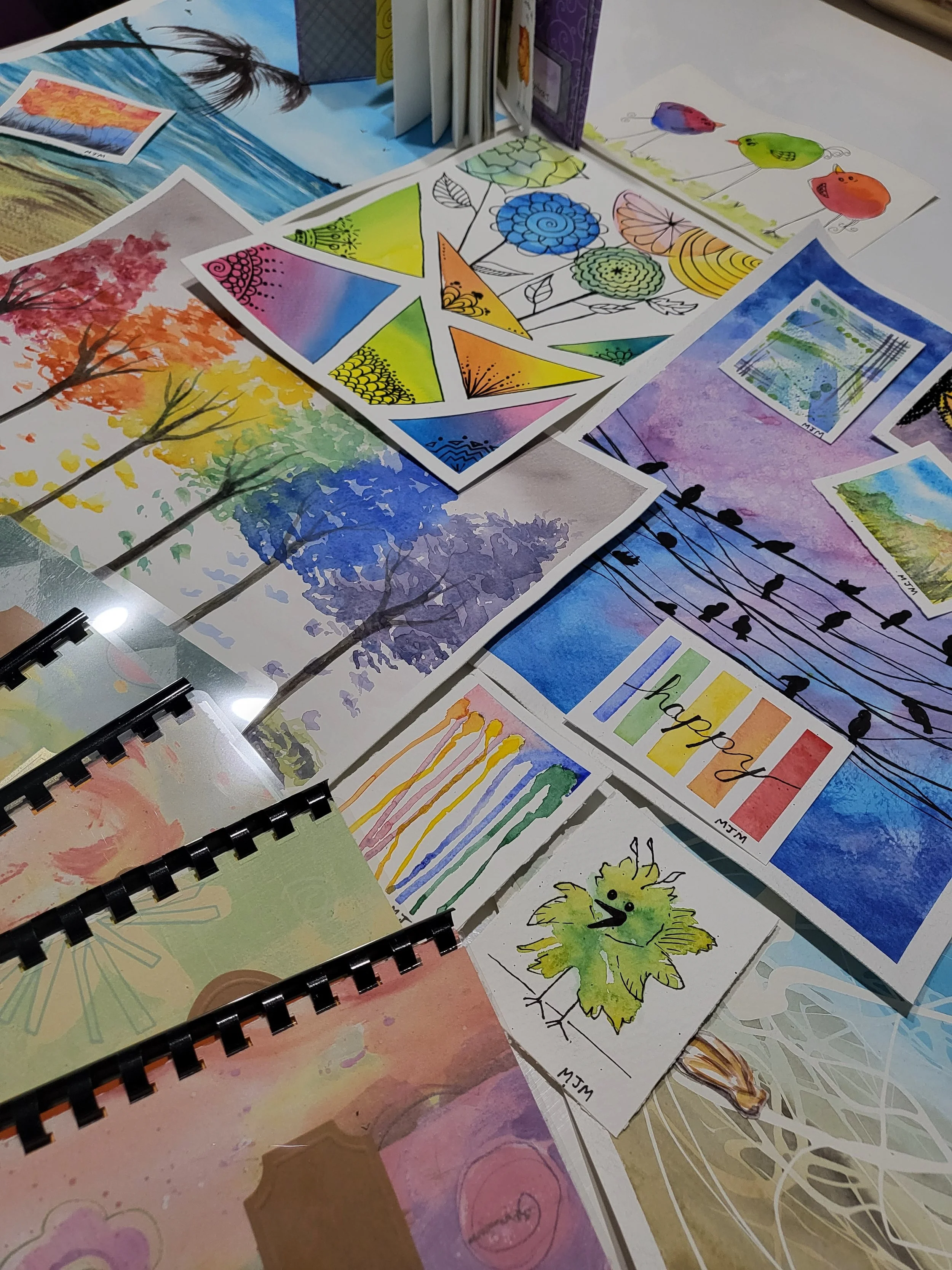 Collection of colorful watercolor paintings and sketches, including trees, flowers, birds, and rainbow-themed designs, spread out on a table.