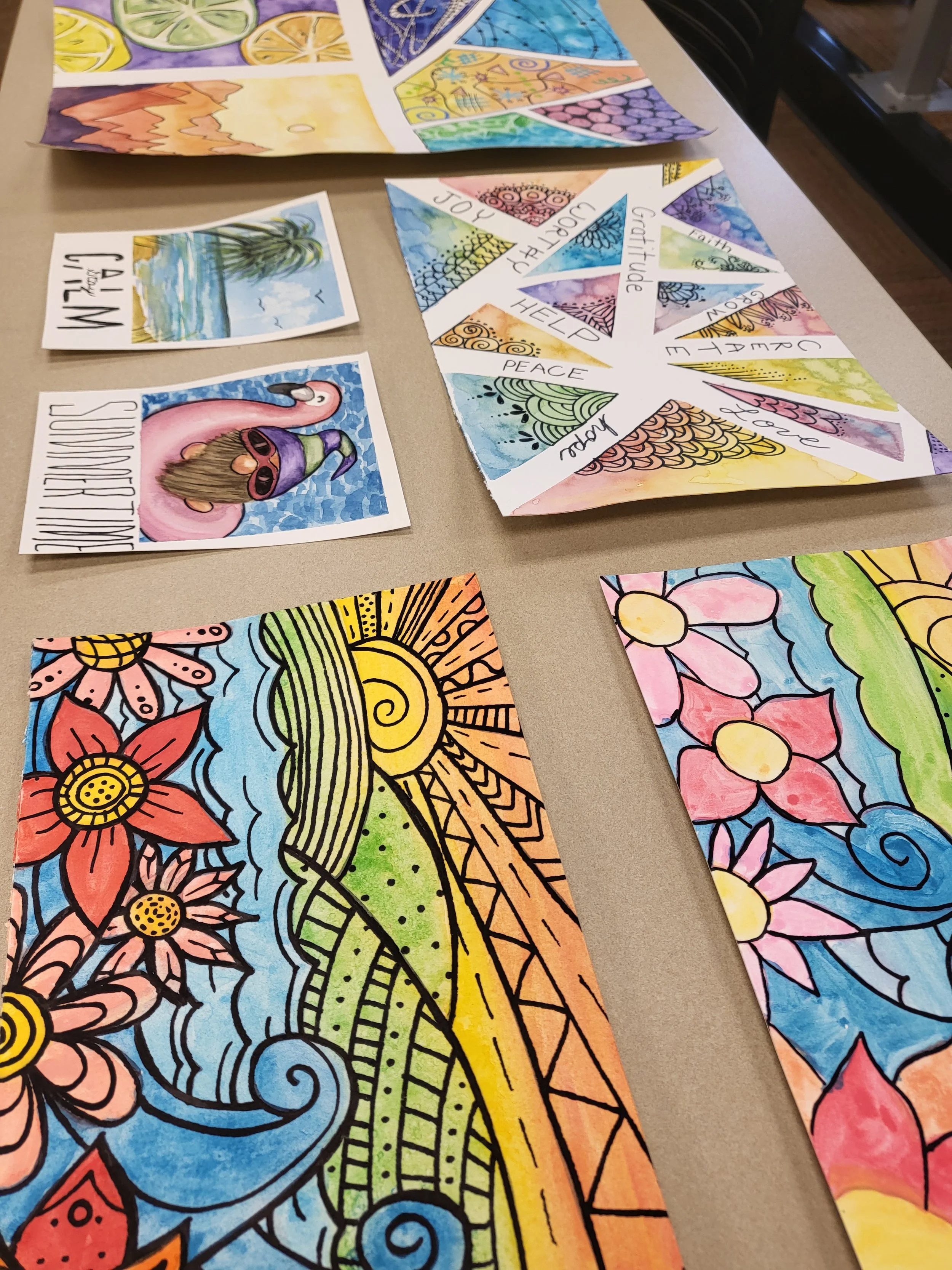 Colorful hand-drawn and painted artwork on paper with floral, landscape, and abstract designs, some including words like 'JOY,' 'PEACE,' and 'HELP.'