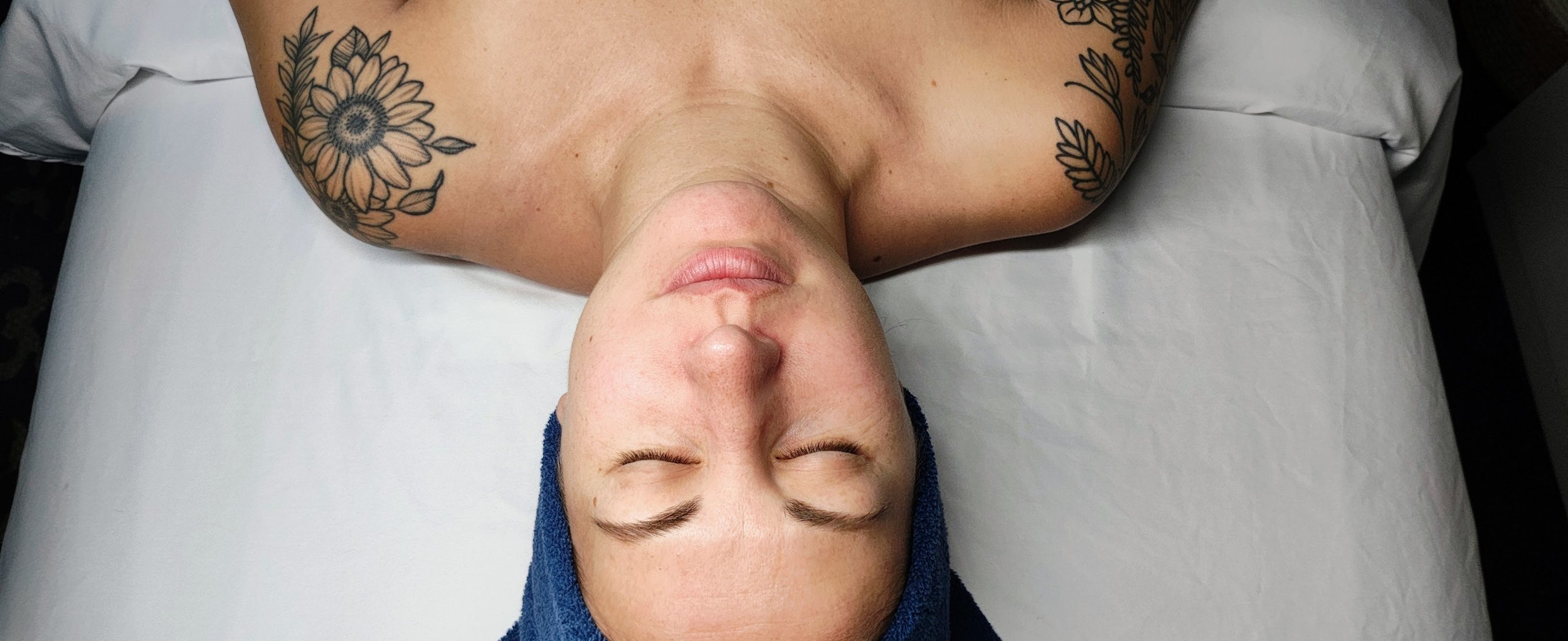 Top-down view of a woman lying on a massage table with a blue towel wrapped around her head, eyes closed, and tattoos of flowers on her shoulder.