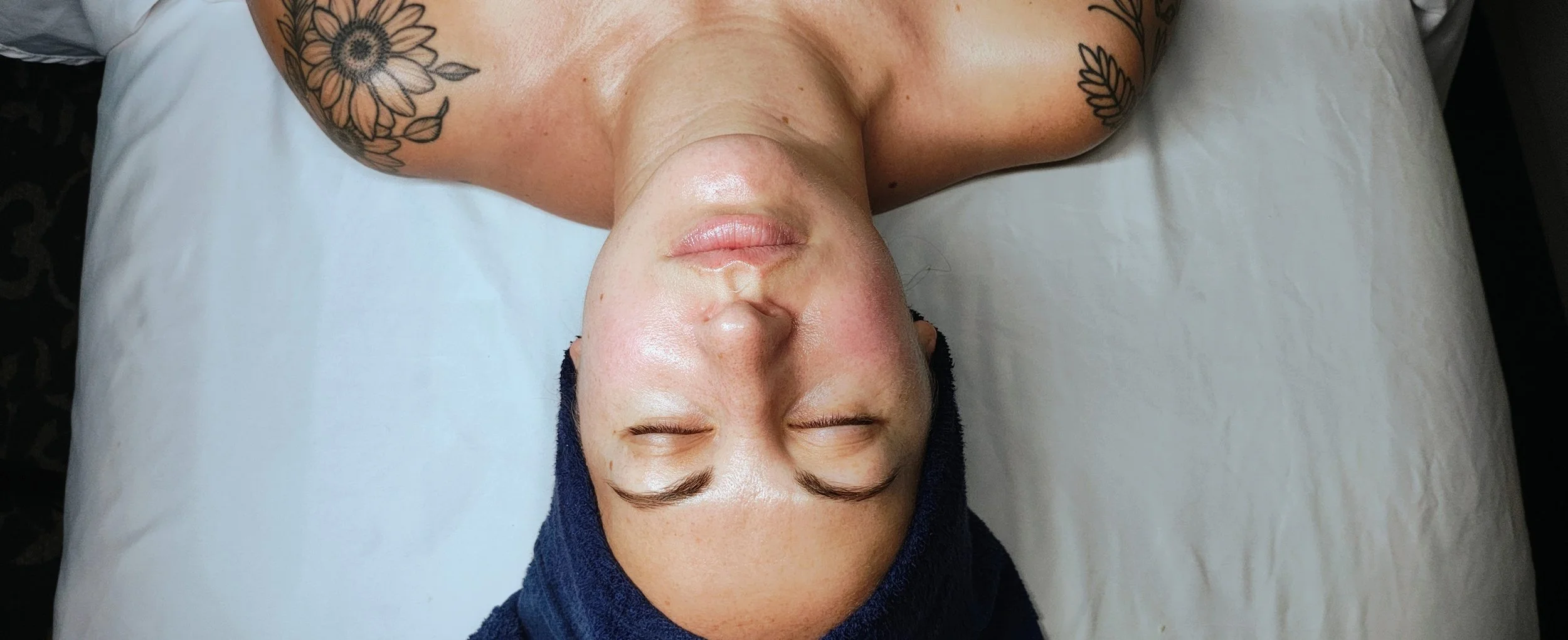 A person lying on a bed with their eyes closed, wearing a black towel on their head and showing tattoos on their shoulders.