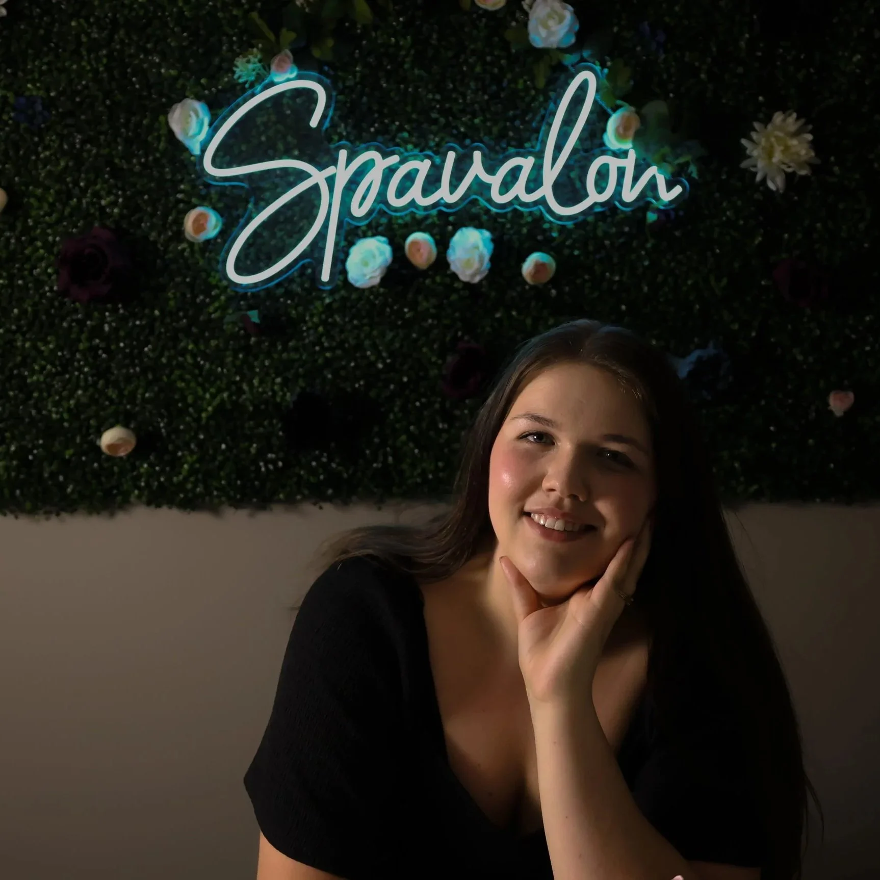 A woman sitting in front of a green hedge wall decorated with flowers and a neon sign that says 'Sparalo' in cursive script. She has long brown hair, is smiling, and resting her chin on her hand.