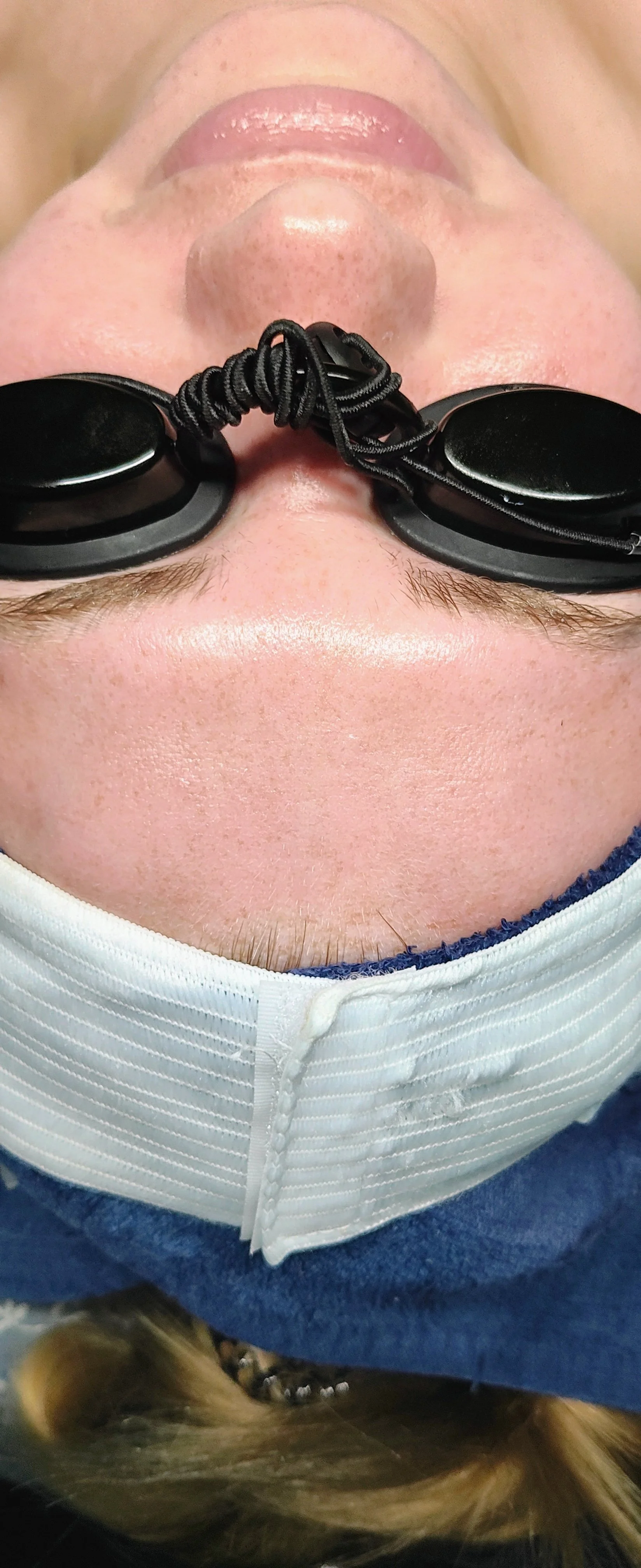 Upside-down image of a person's face wearing black goggles with a cord, a white mask below the nose, and brown hair visible at the bottom.