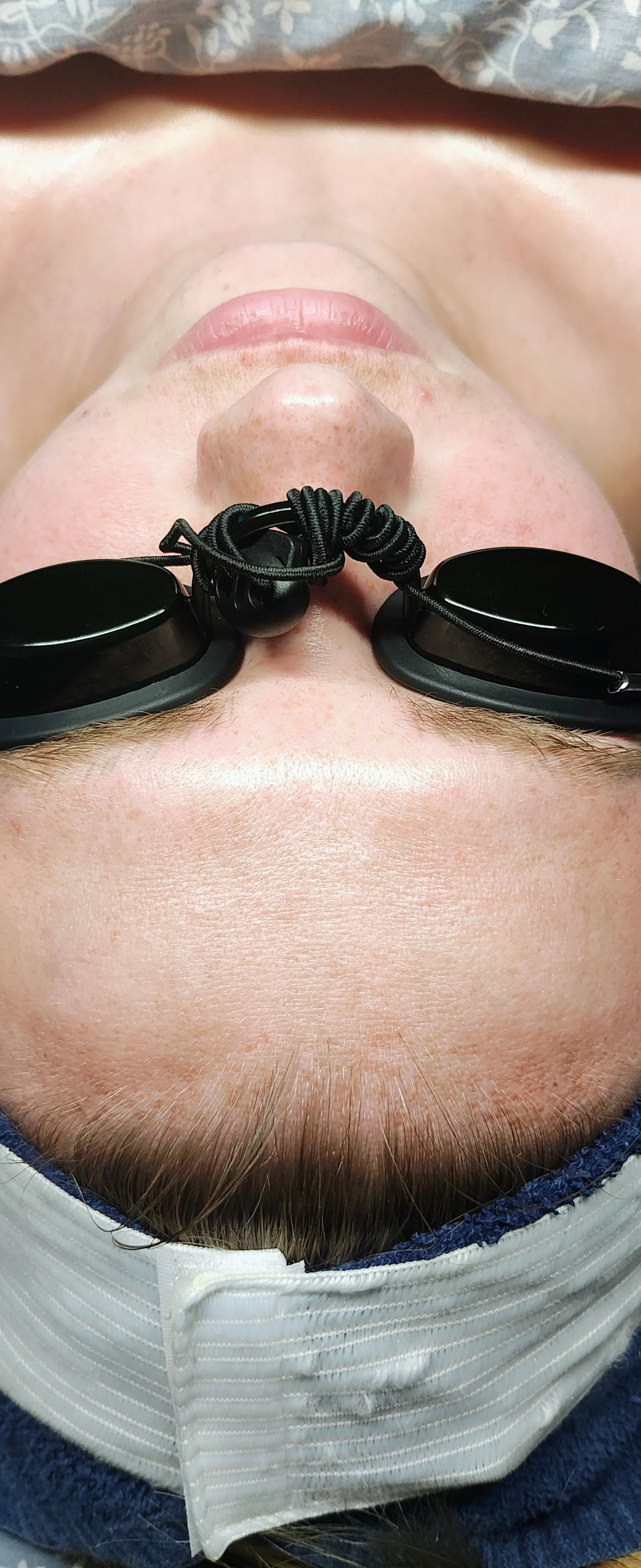 Person lying down, wearing protective goggles and a hair cover, with a towel or cloth over their eyes.