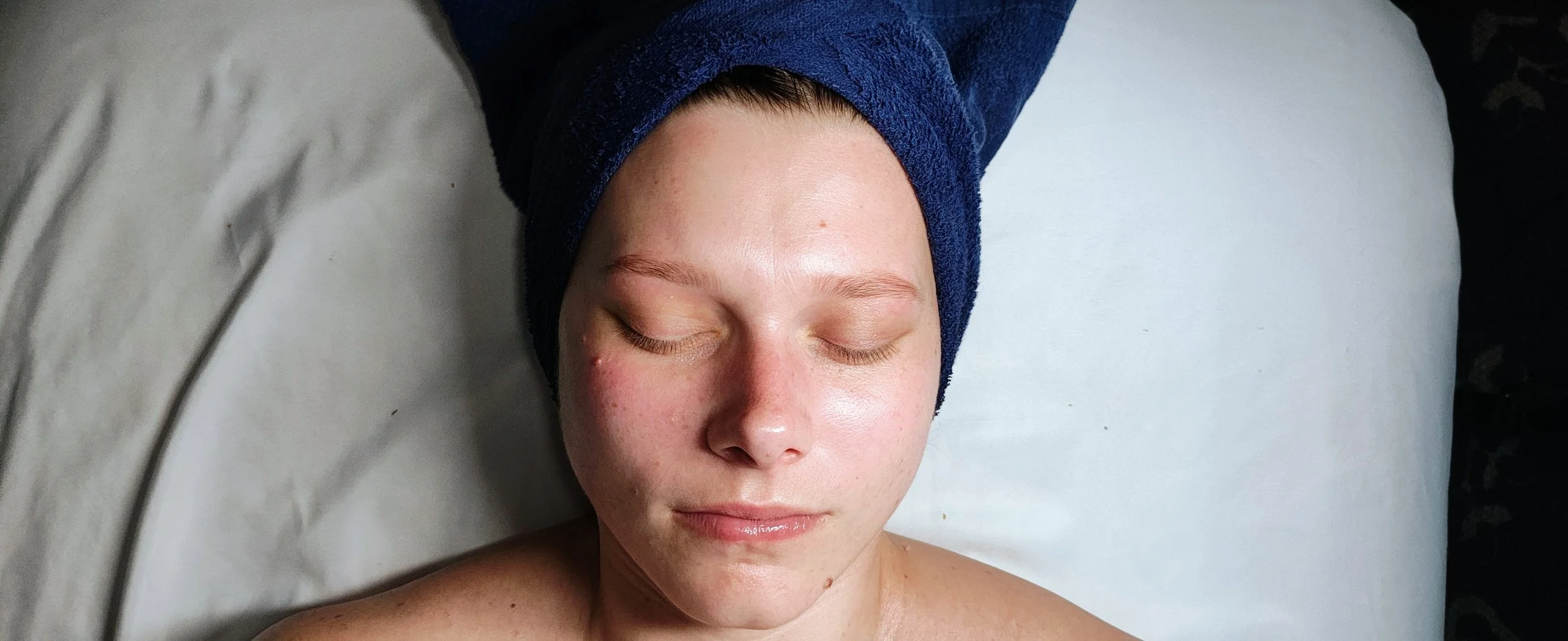 A woman with a towel wrapped around her head, lying down with eyes closed, on a pillow, appearing relaxed after a spa or skincare treatment.