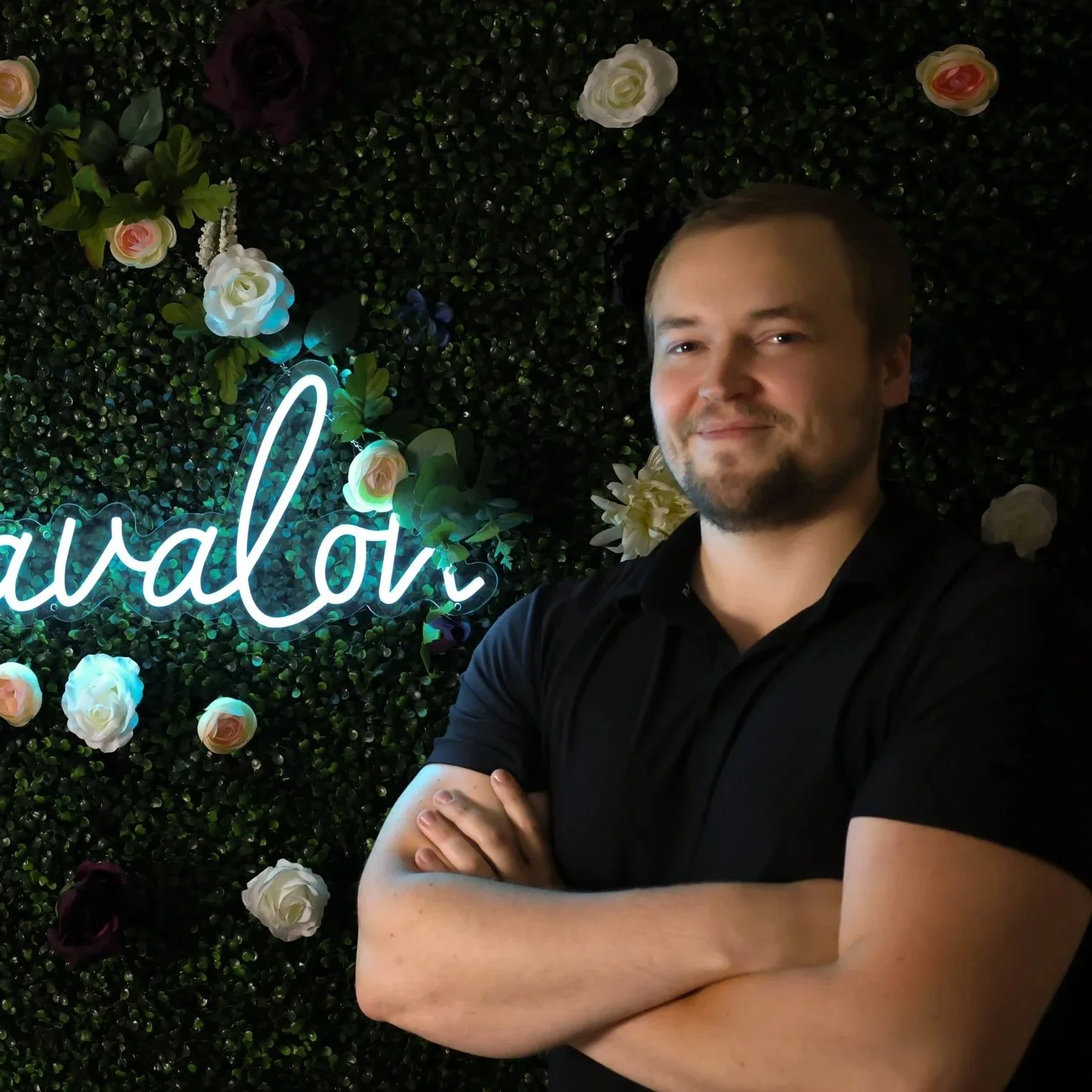 A man with crossed arms standing in front of a decorative wall with artificial flowers and a neon sign that reads 'balov'.