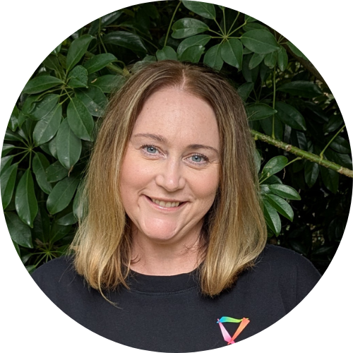 A woman with shoulder-length red hair and blue eyes smiling indoors with a window showing trees in the background.