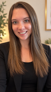 A woman with long brown hair, smiling, wearing a black top and blazer, in an indoor setting with decor and plants in the background.