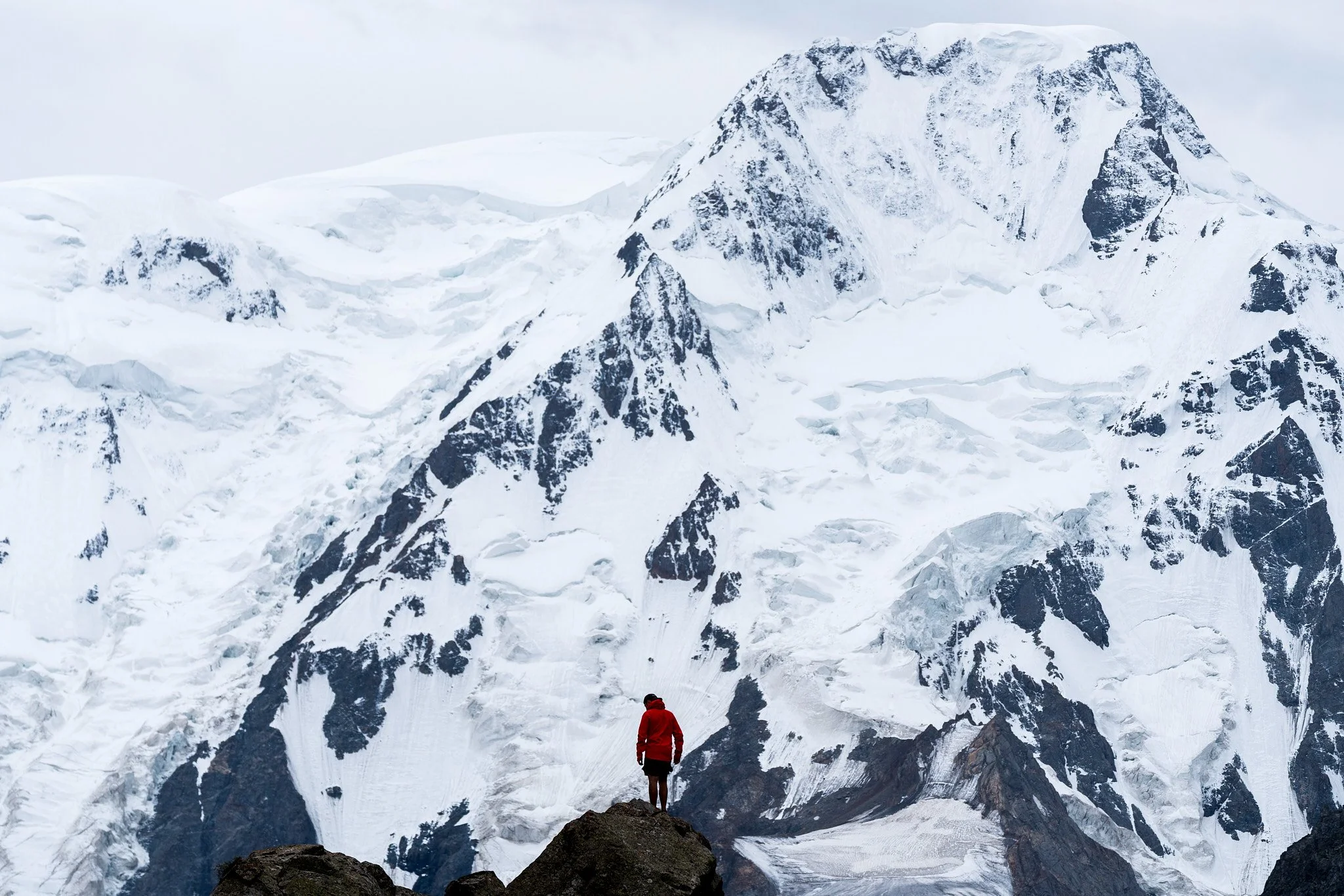 A person in a red jacket standing on a rock overlooking a snowy mountain glacier with high, jagged peaks in the background.