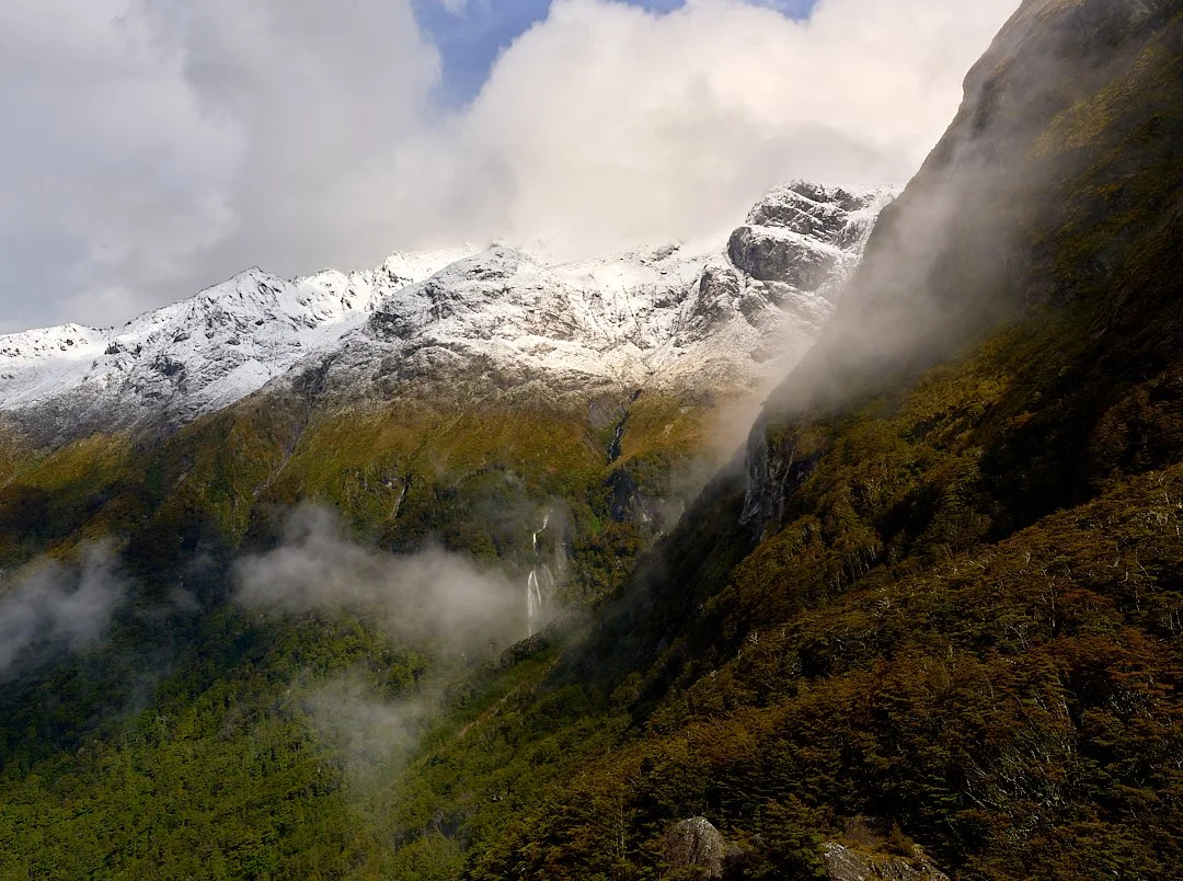 Snow-capped mountains with a forested valley, waterfalls, and clouds in a scenic landscape.