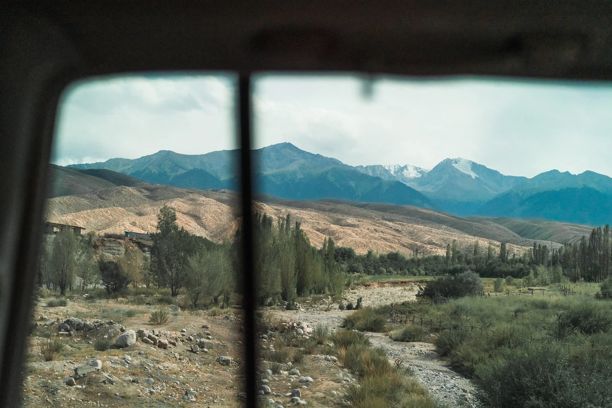 Mountain landscape viewed through a window, with rugged mountains in the distance and a dry, rocky valley with sparse vegetation and trees in the foreground.