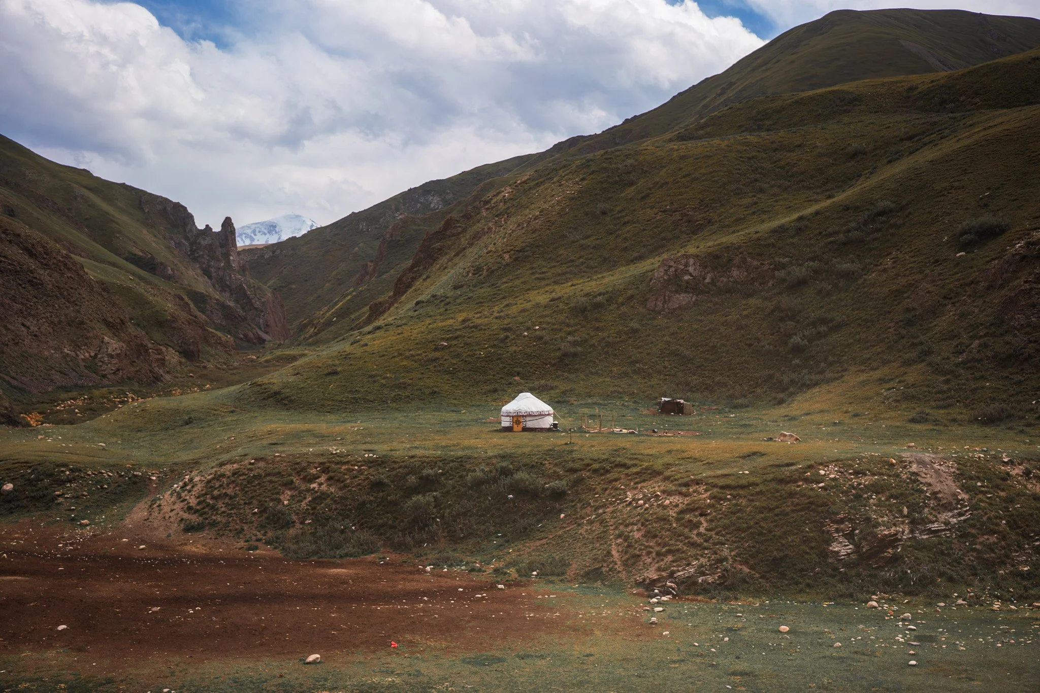 A lush green valley with mountains on either side, a small white yurt in the middle, and a cloudy sky overhead.