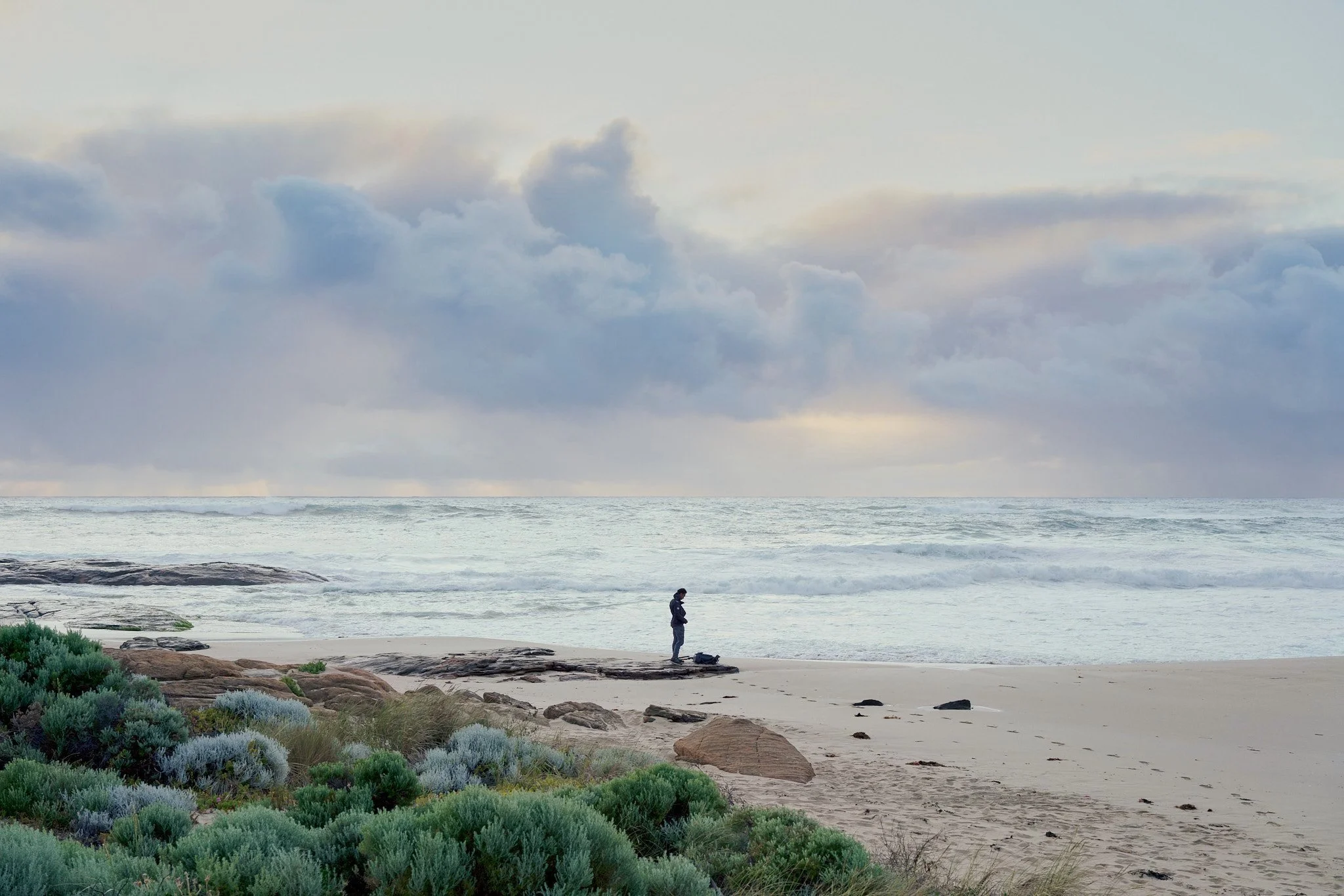 A person standing on a rocky section of a beach, looking down at their phone, with a backpack nearby, surrounded by greenery, ocean waves, and a cloudy sky. Western Australia