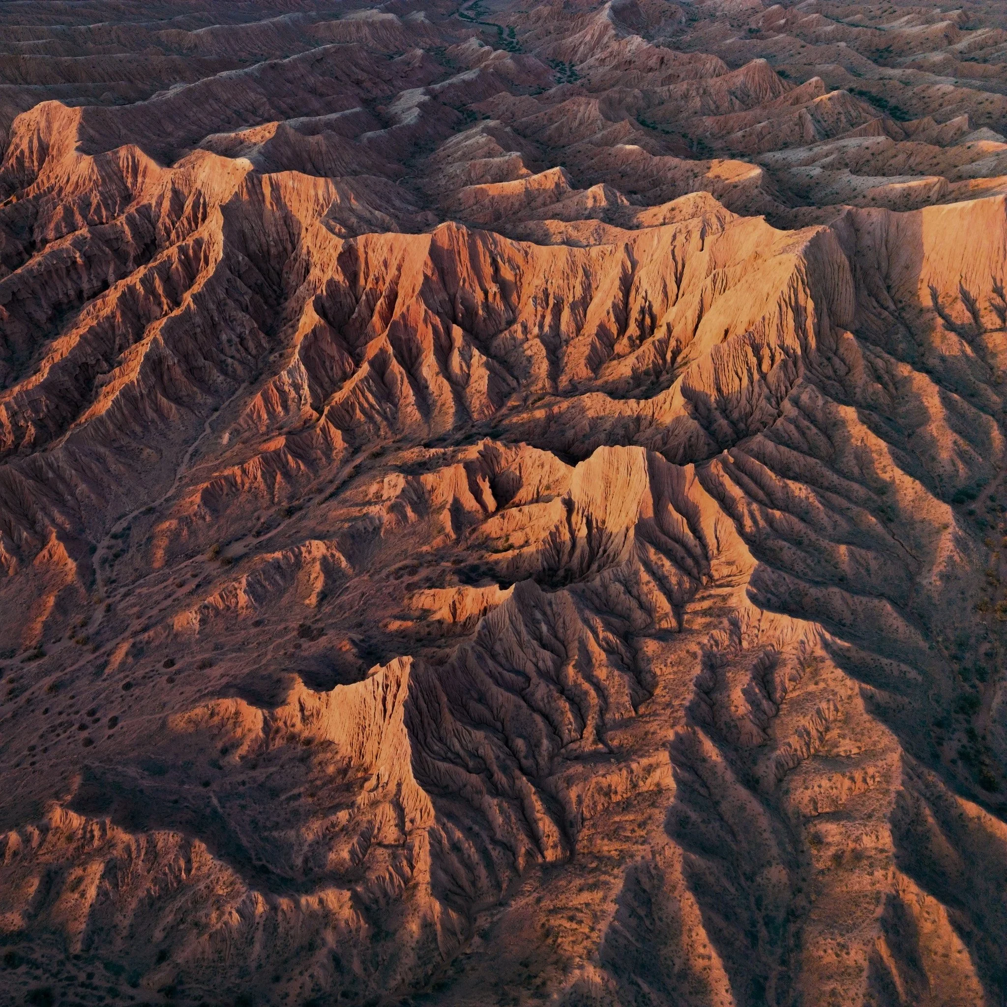 Aerial view of colorful canyon landscape with rugged ridges and deep valleys, illuminated by warm sunlight.