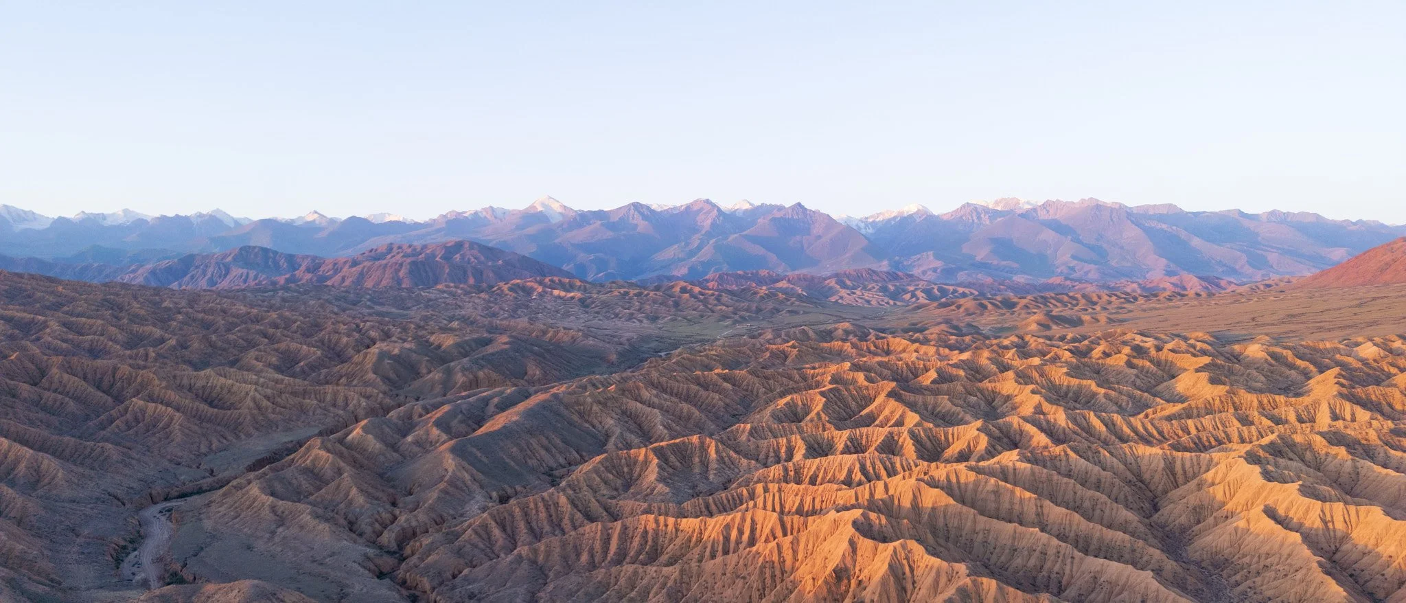 Mountain landscape with rugged brown hills in the foreground and snow-capped peaks in the background under clear sky.