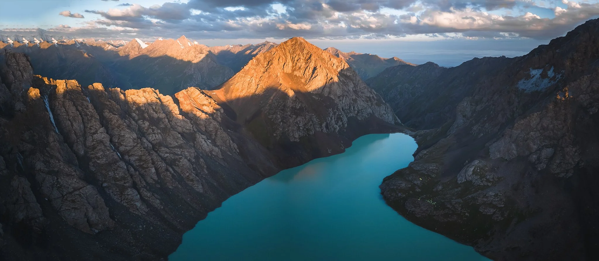 A mountainous landscape at sunset with a turquoise lake in a valley, rugged peaks, snow-capped mountains in the distance, and partly cloudy sky.