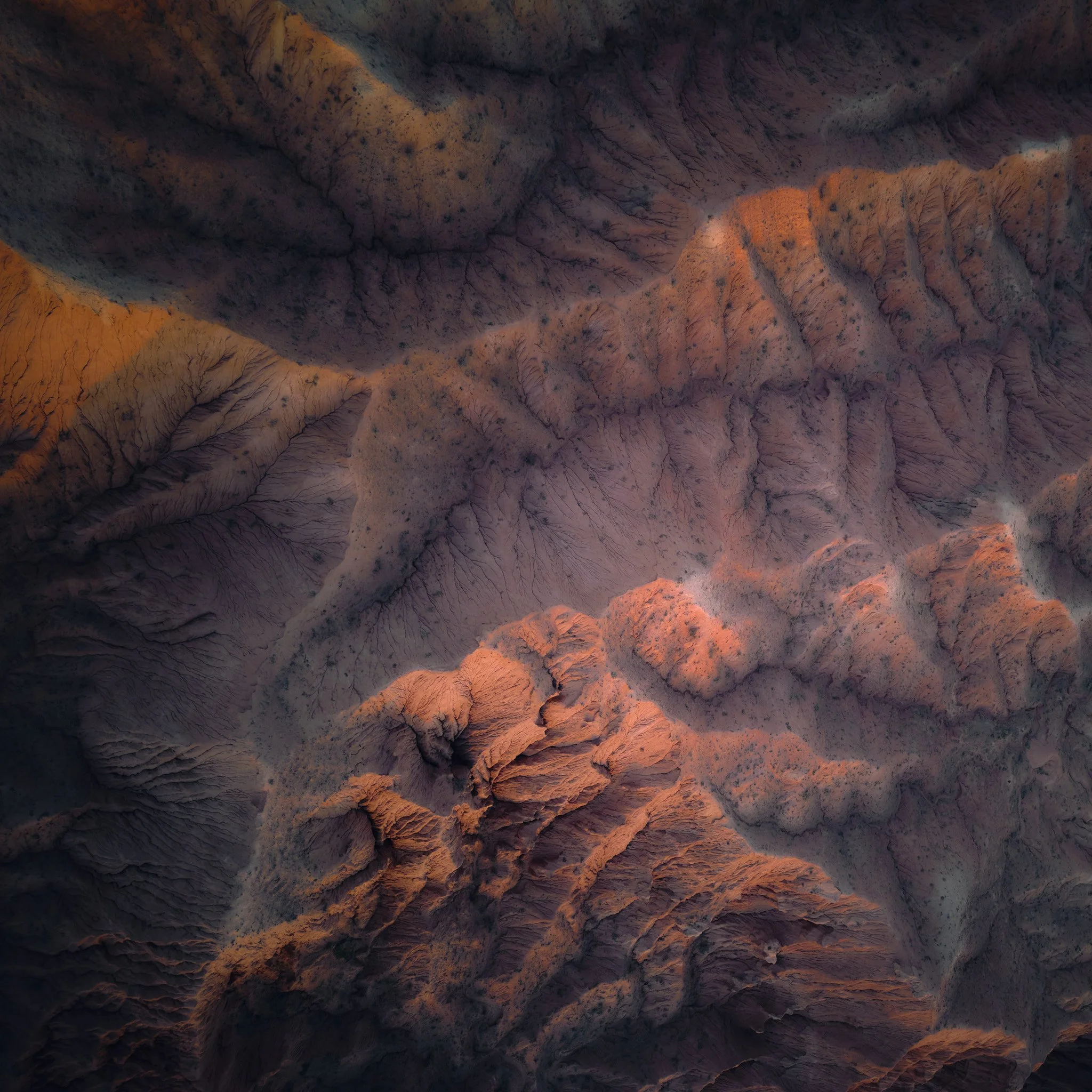 Aerial view of rugged desert mountains with sharp ridges and deep canyons in warm orange and purple hues.