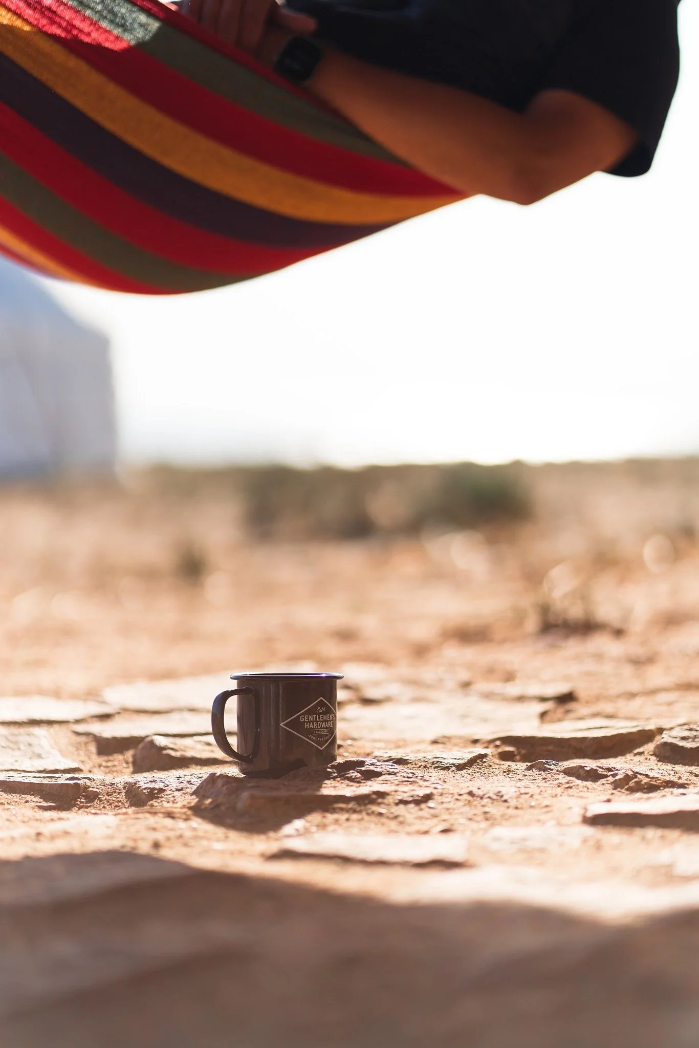A black mug on a rocky surface outdoors, with a colorful striped fabric overhead and a blurred background of desert landscape and sky.