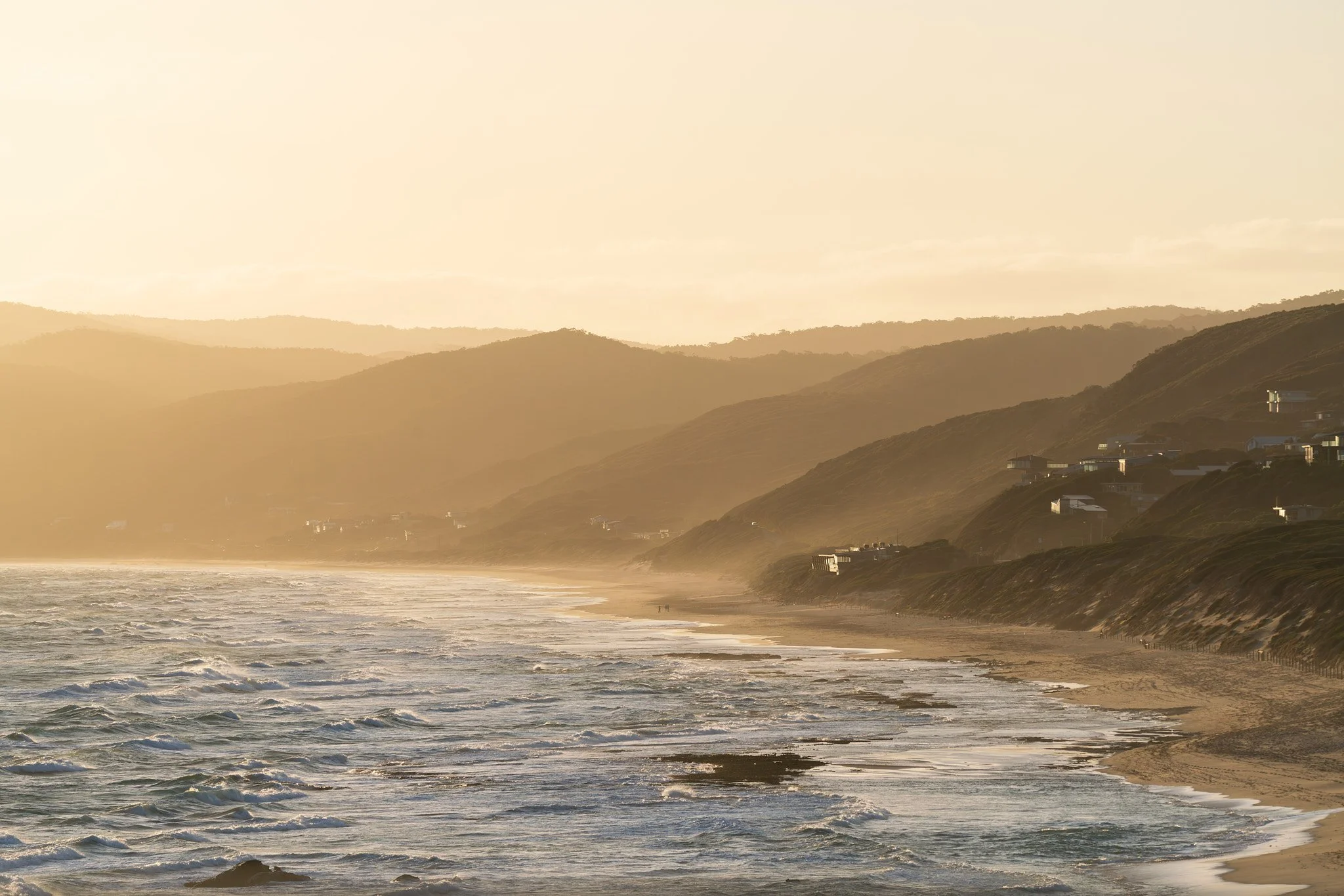 Golden hour at a beach with waves and hills with scattered houses in the background.